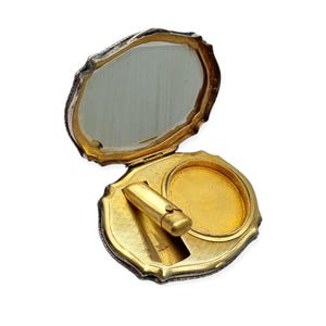May include: An open, scalloped-edge compact case with a silver-toned exterior and a gold-toned interior. The lid features an integrated mirror, while the base contains a circular powder well and a small, cylindrical gold-toned lipstick holder.