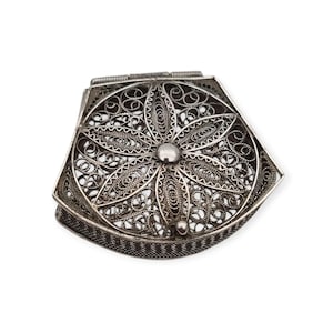 May include: A silver-colored metal box with a hinged lid featuring intricate filigree wirework in a floral pattern. The box has a pentagonal shape with a textured side border and a central circular stud on the top surface of the lid.