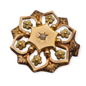 Puede incluir: Un broche dorado con un diseño central en forma de estrella y una sola piedra nacarada. El broche está rodeado de un delicado patrón floral.
