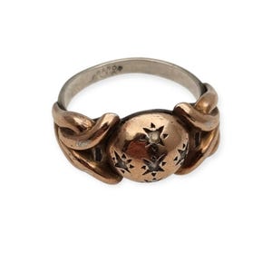 May include: A metallic ring featuring a central rounded element adorned with engraved star patterns. The band displays a twisted, interconnected design in a warm copper tone, while the inner surface of the band shows visible stamped text.