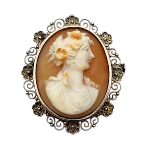 Peut inclure: Broche camée ovale représentant un profil de femme sculpté avec des fleurs dans les cheveux. Le camée est serti dans un cadre filigrané argenté avec des détails floraux. Le camée est de couleur orange clair.