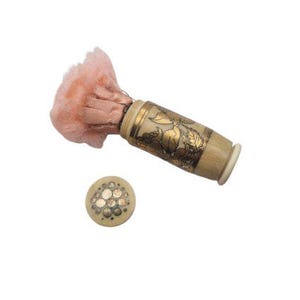 May include: An antique powder brush with a peach-colored puff and a decorative cylindrical handle. The handle is cream-colored with gold floral designs. A small, round, cream-colored lid with gold accents is also present.