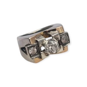 May include: A two-tone metal ring featuring a wide, polished band with a central, larger round clear stone flanked by two smaller round clear stones, each set within a square-shaped frame integrated into the band's design.