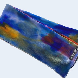 May include: A hand-dyed scarf in shades of blue, orange, and purple. The scarf has a watercolor effect with a label that reads "DIANA HAND Handmade Textiles". The scarf is folded and appears to be made of a soft, flowing fabric.