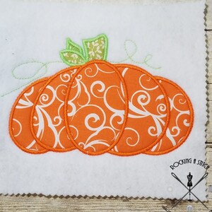 Op de afbeelding: Een wit vierkant van vilt met een oranje pompoen met een groene steel en witte swirls. De tekst "ROCKING A STITCH" staat in de rechterbenedenhoek van de afbeelding.