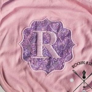 May include: Pink baby bib with a white embroidered letter R in a purple paisley design. The bib has the text "#953 FRAME TWO" and "ROCKING A STITCH" on it.