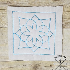 May include: A white fabric square with a blue embroidered geometric design. The design features a flower-like shape with overlapping circles and curved lines. The fabric has a subtle texture and the embroidery is precise and detailed. The bottom right corner of the fabric has the text "Rocking Stitch" and a small logo with a sewing needle and thread.