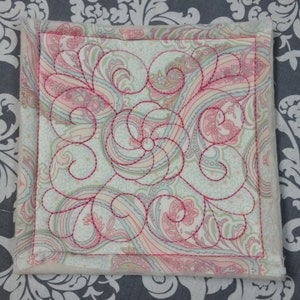 May include: A square coaster with a white background and a pink paisley pattern. The coaster is quilted with a pink thread in a swirling pattern.