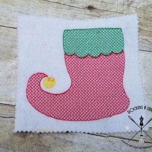 Puede incluir: Zapato de elfo rojo y verde con una campanita amarilla. El zapato está bordado con un patrón de puntadas.