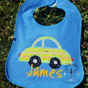 May include: A blue baby bib with a green and yellow car design and the name "James" embroidered on it. The bib has a white trim and the text "Rockin' a Stitch" in black with a needle and thread logo.
