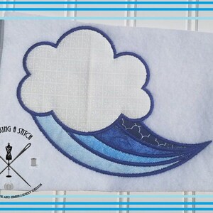 May include: A blue and white embroidered design of a cloud with a blue wave coming out of it. The design is on a white fabric background. The text "Rocking a Stitch" is in the lower left corner of the image.