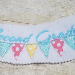 Peut inclure: Bannière en feutre brodée avec le texte "Second Grade" en bleu, avec une chaîne de fanions colorés et un dessin de crayon.