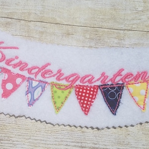 Peut inclure: Une bannière en feutre blanc avec le mot "Kindergarten" brodé en cursive rose. La bannière est décorée d'une rangée de huit triangles en tissu coloré, chacun avec un motif différent.