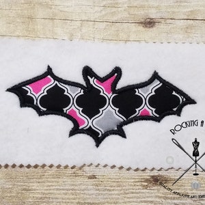 Op de afbeelding: Zwarte en roze gepatroneerde vleermuis-applicatie met een zwarte omtrek. De vleermuis bevindt zich op een witte achtergrond. "Rocking a Stitch" staat in zwarte tekst onder de vleermuis.