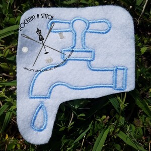 Puede incluir: Diseño bordado en azul de un grifo con una gota de agua, sobre un fondo de tela blanca. El diseño incluye el texto "Rocking a Stitch" y "Quality Applique and Embroidery Design".