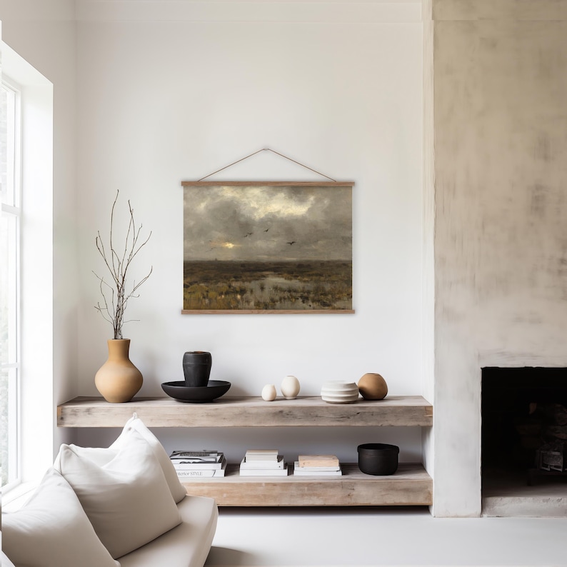 A living room featuring a large hanging tapestry featuring a dark and moody landscape painting of a marsh, the sun trying to come out from behind the clouds and birds flying overhead.  The tapestry it strimmed at the top and bottom with oak battons and an adjustable vegan friendly cord so you cna hang the tapestry at the right height