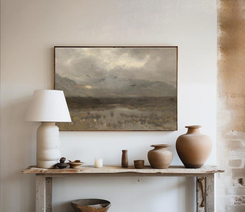 A styled hallway console with a large hanging painting featuring a dark and moody landscape painting of a marsh, the sun trying to come out from behind the clouds and birds flying overhead. Accessories the hallway table.