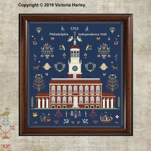 May include: A cross-stitch sampler depicting Independence Hall in Philadelphia, Pennsylvania. The building is red brick with white trim and a clock tower. The sampler is framed in brown wood and has a blue background with various floral and animal motifs. The text "1753", "Philadelphia", and "Independence Hall" is included in the sampler.
