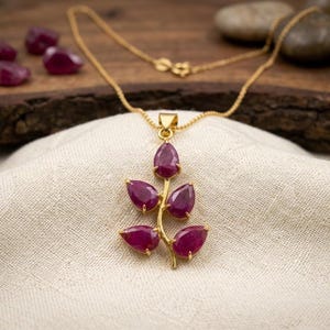 Peut inclure: Un collier en or avec un pendentif en forme de branche orné de cinq pierres précieuses rubis en forme de poire, d'un rouge rosé profond. Le pendentif est suspendu à une fine chaîne en or, évoquant une tige naturelle avec des feuilles.