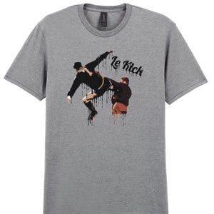 Puede incluir: Camiseta gris con un gráfico de dos figuras en una pose de acción de fútbol, con las palabras "Le Kick" en una fuente goteante. Las figuras son en negro y marrón, con gotas negras. La camiseta tiene cuello redondo.