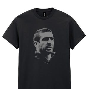 Puede incluir: Camiseta negra con un retrato en escala de grises de un hombre. El retrato está centrado en la camiseta y muestra al hombre desde el pecho hacia arriba. La camiseta tiene cuello redondo y mangas cortas.