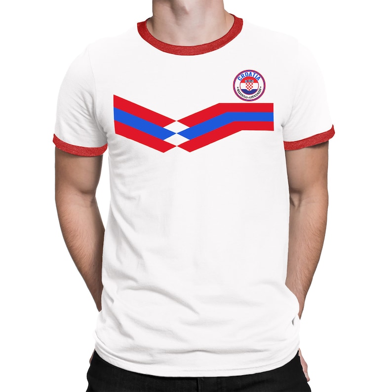 May include: White t-shirt with a red and blue chevron design and a red ringer neck. The shirt has a circular patch with the text "Croatia" and a red, white, and blue checkered pattern.