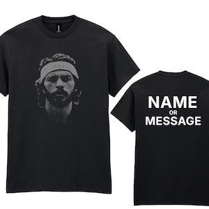 May include: Black t-shirt featuring a grayscale portrait of a man wearing a headband. The back of the shirt displays the text "NAME OR MESSAGE" in white block letters. The shirt is a crew neck style.