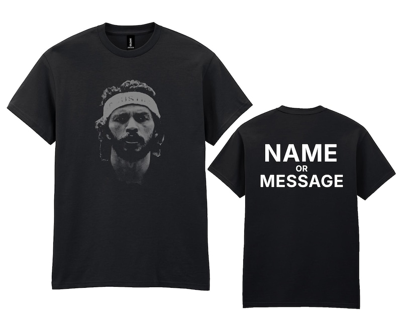 May include: Black t-shirt featuring a grayscale portrait of a man wearing a headband. The back of the shirt displays the text "NAME OR MESSAGE" in white block letters. The shirt is a crew neck style.