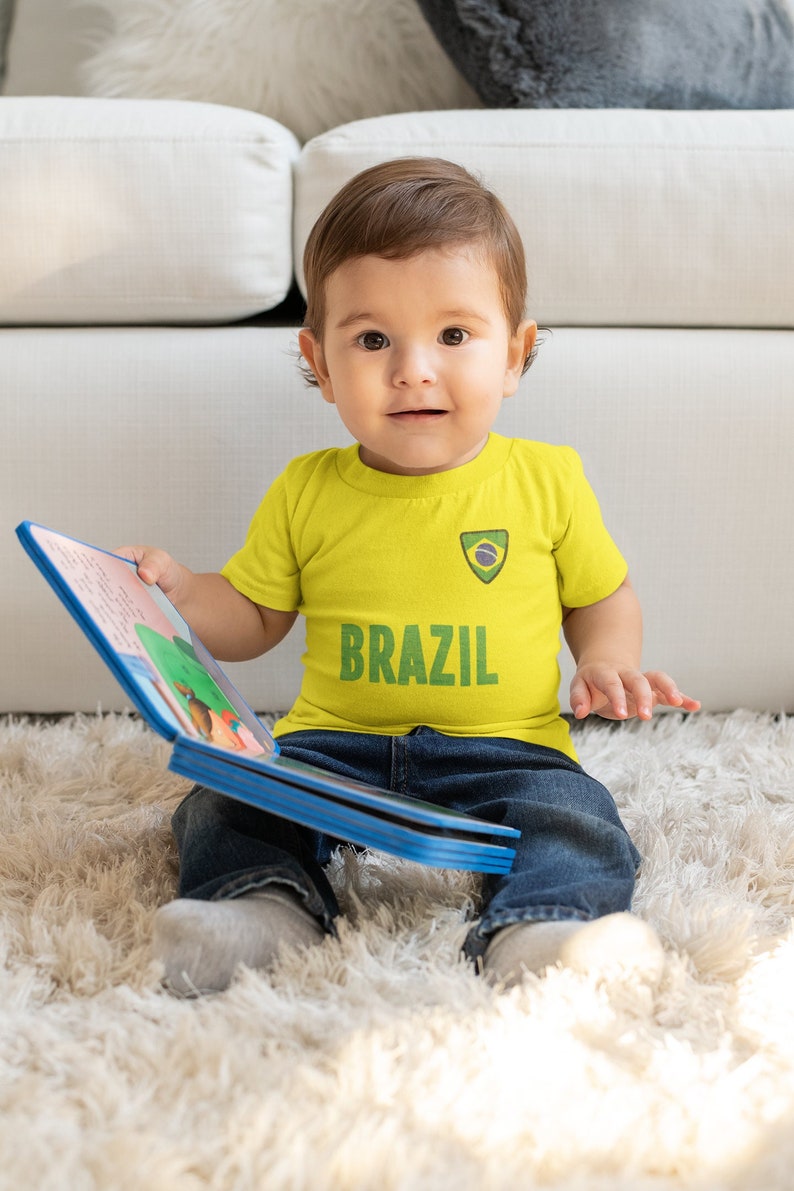 May include: A young child wearing a yellow t-shirt with a green and blue Brazilian flag patch and the word "BRAZIL" printed on it. The child is sitting on a white fluffy rug and holding a colorful book.