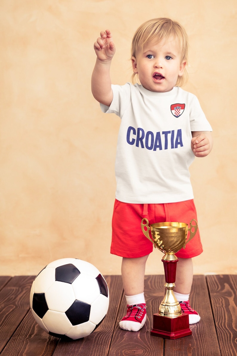 May include: A young child wearing a white soccer jersey with the word "CROATIA" and the Croatian flag on it. The child is holding a gold trophy and a black and white soccer ball. The child is standing on a wooden floor.
