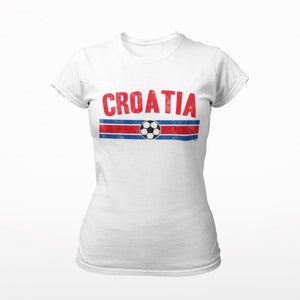 May include: White t-shirt with a red and blue stripe design and a soccer ball graphic. The word "CROATIA" is printed in red letters above the stripe.