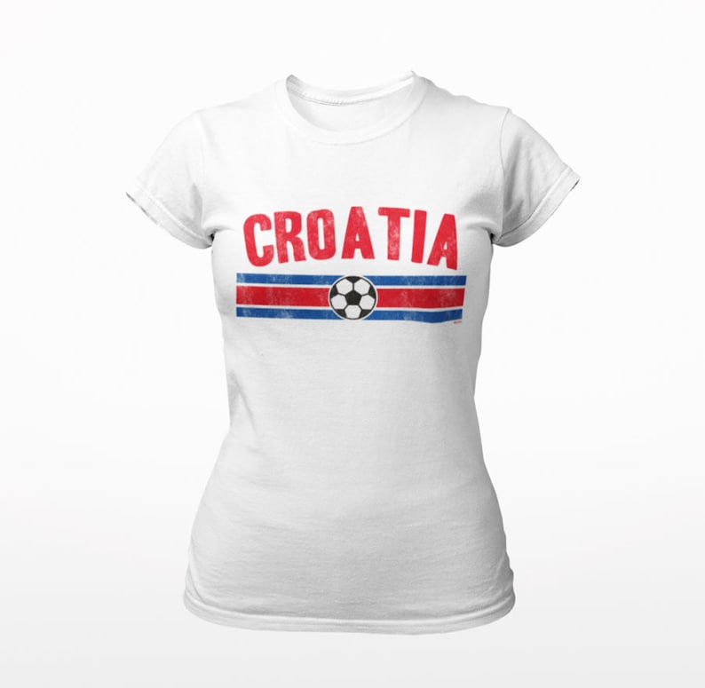 May include: White t-shirt with a red and blue stripe design and a soccer ball graphic. The word "CROATIA" is printed in red letters above the stripe.