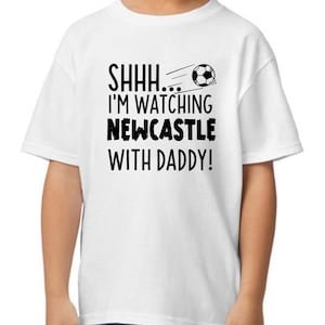 Peut inclure: T-shirt blanc avec du texte noir qui dit "SHHH... I'M WATCHING NEWCASTLE WITH DADDY!" et un graphique de ballon de football noir et blanc.