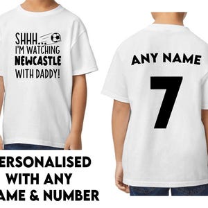Peut inclure: Deux t-shirts blancs, l'un avec le texte "SHHH... I'M WATCHING NEWCASTLE WITH DADDY!" et un graphique de ballon de football, et l'autre avec le texte "ANY NAME" et le num&eacute;ro "7" imprim&eacute; au dos.