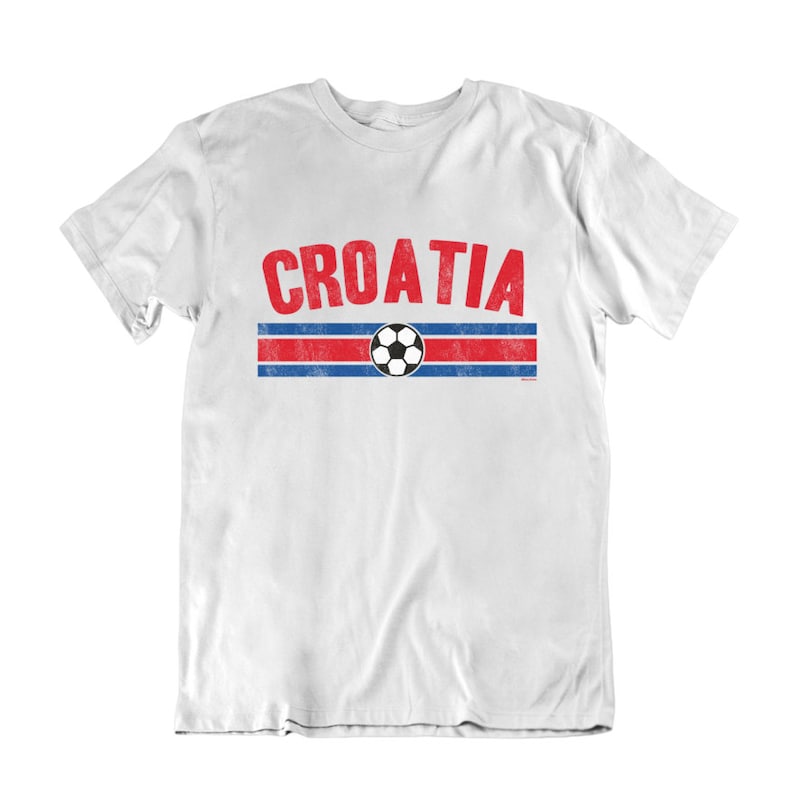 May include: White t-shirt with a red and blue striped design and a soccer ball. The word "CROATIA" is printed in red letters above the stripes.