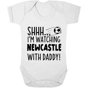 Peut inclure: Combinaison blanche pour b&eacute;b&eacute; avec un texte noir qui dit "Chut... Je regarde Newcastle avec papa !" et un dessin de ballon de football noir et blanc.