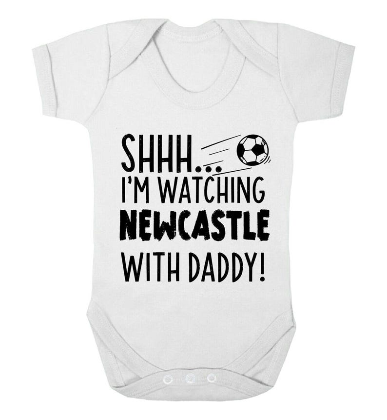 Peut inclure: Combinaison blanche pour b&eacute;b&eacute; avec un texte noir qui dit "Chut... Je regarde Newcastle avec papa !" et un dessin de ballon de football noir et blanc.