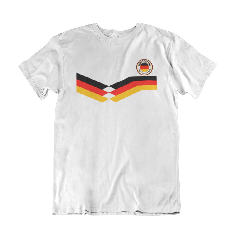 May include: White t-shirt with a black, yellow and red German flag design on the front. The flag design is in the shape of a chevron. The shirt also has a small circular patch with the word "Deutschland" in black text on a white background.