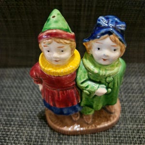 Op de afbeelding: Een keramische zout- en peperstelset met twee figuren die samen op een bruine basis staan. Het linkerfiguur draagt een groene puntmuts, een gele kraag en een rode jas. Het rechterfiguur draagt een blauwe hoed en een groene jas met knopen.