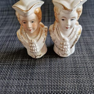 May include: A pair of white ceramic salt and pepper shakers shaped like colonial-era busts. Each figure wears a tricorne hat and period clothing with gold-painted accents. The faces feature painted eyes, lips, and hair, with a single hole on top.