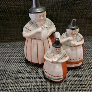 May include: A set of three ceramic figurines depicting figures in traditional attire, each wearing a tall black hat. The figures feature white aprons with red or green stripes, light green shawls, and painted facial details, arranged in a group.