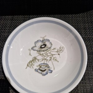 May include: A round white ceramic bowl featuring a light blue band around the inner rim and a central botanical illustration of two blue and green anemone flowers with detailed dark centers and delicate stems and leaves on the white surface.