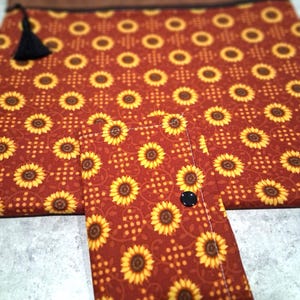 May include: A rectangular fabric pouch and a matching smaller wallet, both featuring a repeating sunflower pattern on a dark red background. The pouch has a brown top border, a black zipper, and a black tassel. The wallet has a black snap button.