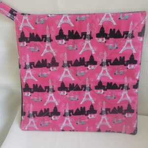 May include: Pink square pot holder with a repeating pattern of the Eiffel Tower, cityscapes, and scooters. The pot holder has a loop for hanging and is quilted with a diagonal stitch pattern.