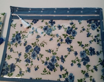May include: A rectangular zippered pouch featuring a clear front panel with a blue and green floral pattern. The pouch has a blue fabric border with a small white cross print and a white zipper pull with a matching white tassel attached to the side.