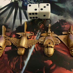 May include: Four gold-colored game pieces with an airplane design, and two white dice with black dots. The game pieces have a detailed, stylized design. The background is a dark, patterned surface.
