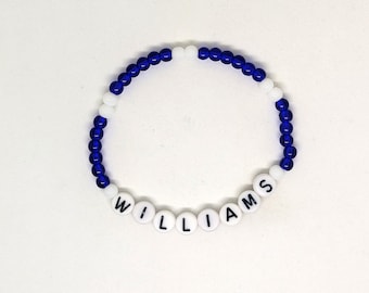 May include: A beaded bracelet with the name "WILLIAMS" spelled out in white letter beads. The bracelet features blue and white beads, creating a contrasting color scheme. The bracelet is displayed on a white surface.