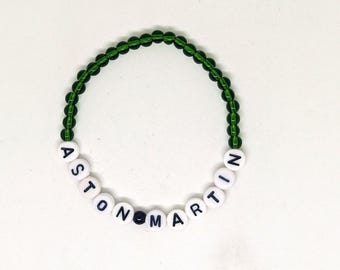 May include: A beaded bracelet with the name "ASTON MARTIN" spelled out in white letter beads. The letters are on a black background. The bracelet's band is made of green beads.