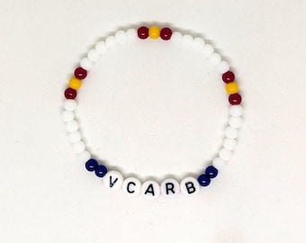 Könnte beinhalten: Weißes Perlenarmband mit dem Wort "VCARB" in weißen Buchstabenperlen. Das Armband hat rote, gelbe und blaue Akzentperlen.