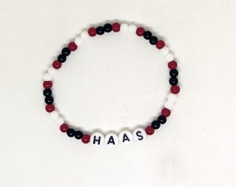 May include: A beaded bracelet with the word "HAAS" spelled out in white letter beads. The bracelet features alternating black, red, and white beads. The bracelet is on a white background.